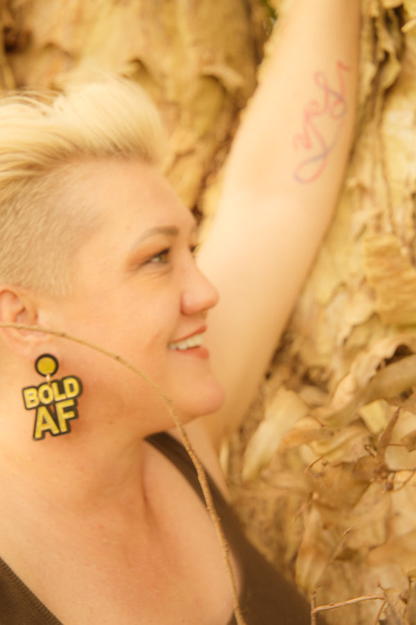 Doodad and Fandango designed earrings on a model depicting the words BOLD AF, in gold and black.