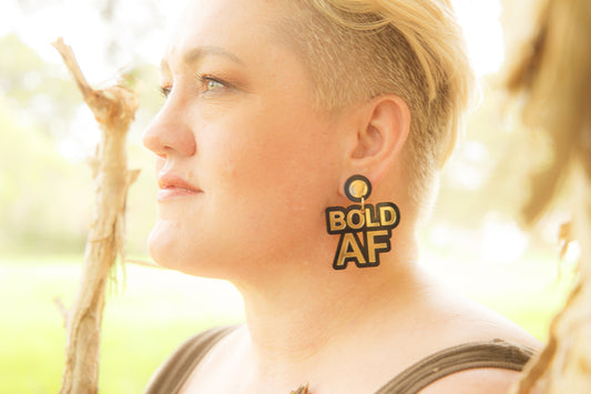 Doodad and Fandango designed earrings on a model depicting the words BOLD AF, in gold and black.
